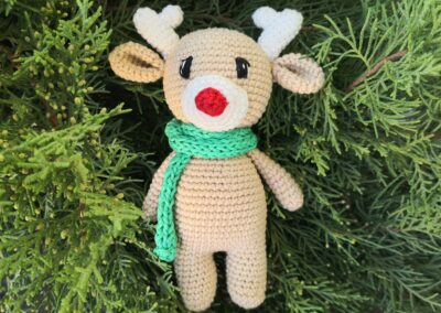 Teljesen elkészült horgolt rénszarvas figura piros orral, sállal és agancsokkal, zöld háttér előtt