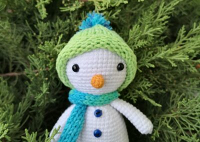 Kézzel horgolt hóember figura sapkában és sálban, fenyőágak előtt fotózva