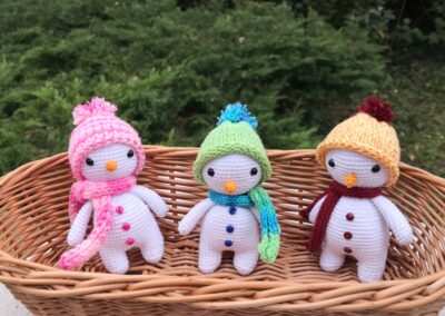 Három kézzel horgolt hóember különböző színű sapkában és sálban, fonott kosárban ülve