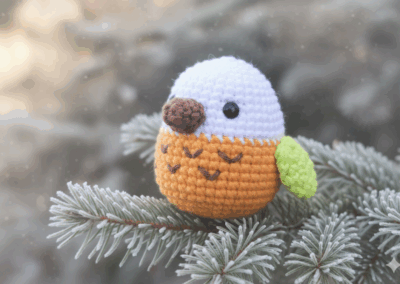 Horgolt madárka karácsonyi fenyőágon – aranyos amigurumi dísz narancs testtel és zöld szárnnyal.