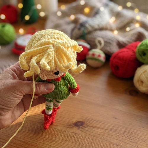 Karácsonyi amigurumi manó felülről, sárga horgolt hajjal, ünnepi fonalakkal a háttérben.