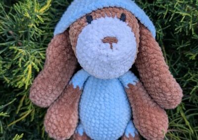 Pizsamás amigurumi kutyus sapkában, puha fonalból horgolva, zöld háttér előtt.